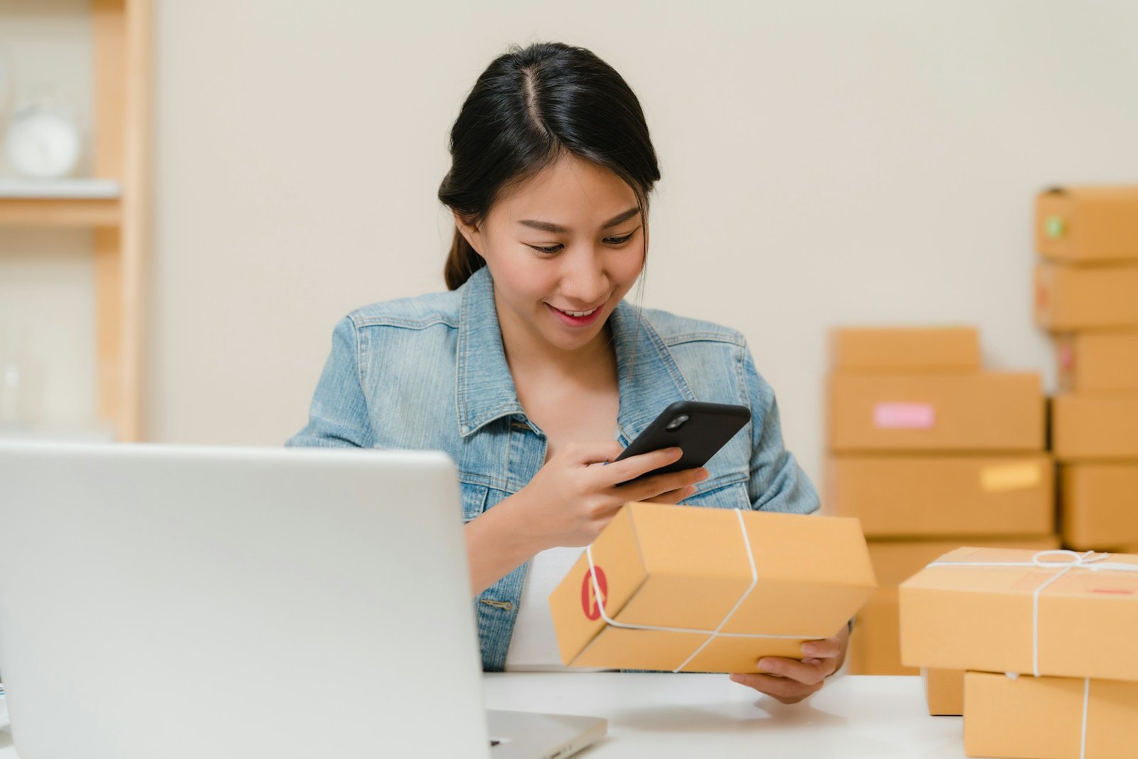 Woman checking a package with her phone near a laptop and boxes, representing economic nexus for online sellers