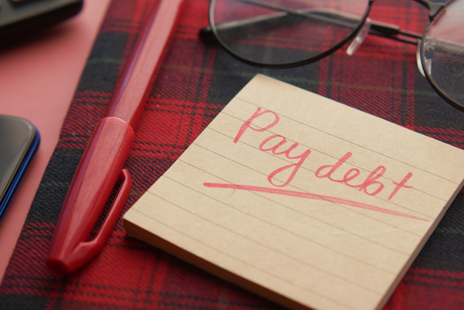 Handwritten note reminding to pay a debt next to a pen and glasses
